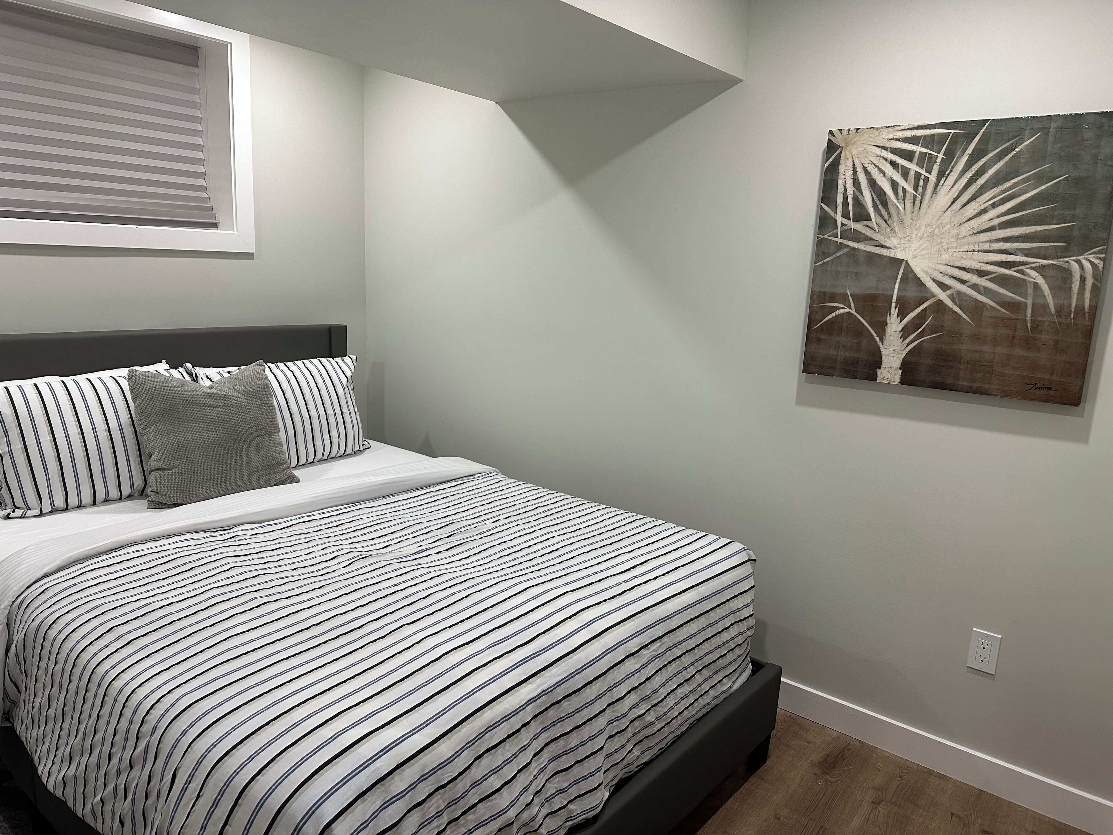 A bedroom with a bed, a window with blinds, a wall-mounted painting, and a power outlet.
