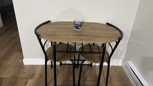 A corner room with a wooden table and two chairs, a small glass container on the table, and a white wall. - Bethel Place - brand new suite in Evergreen! (Saskatoon)
