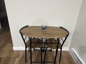Een hoekkamer met een houten tafel en twee stoelen, een klein glazen vaasje op de tafel, en een witte muur.