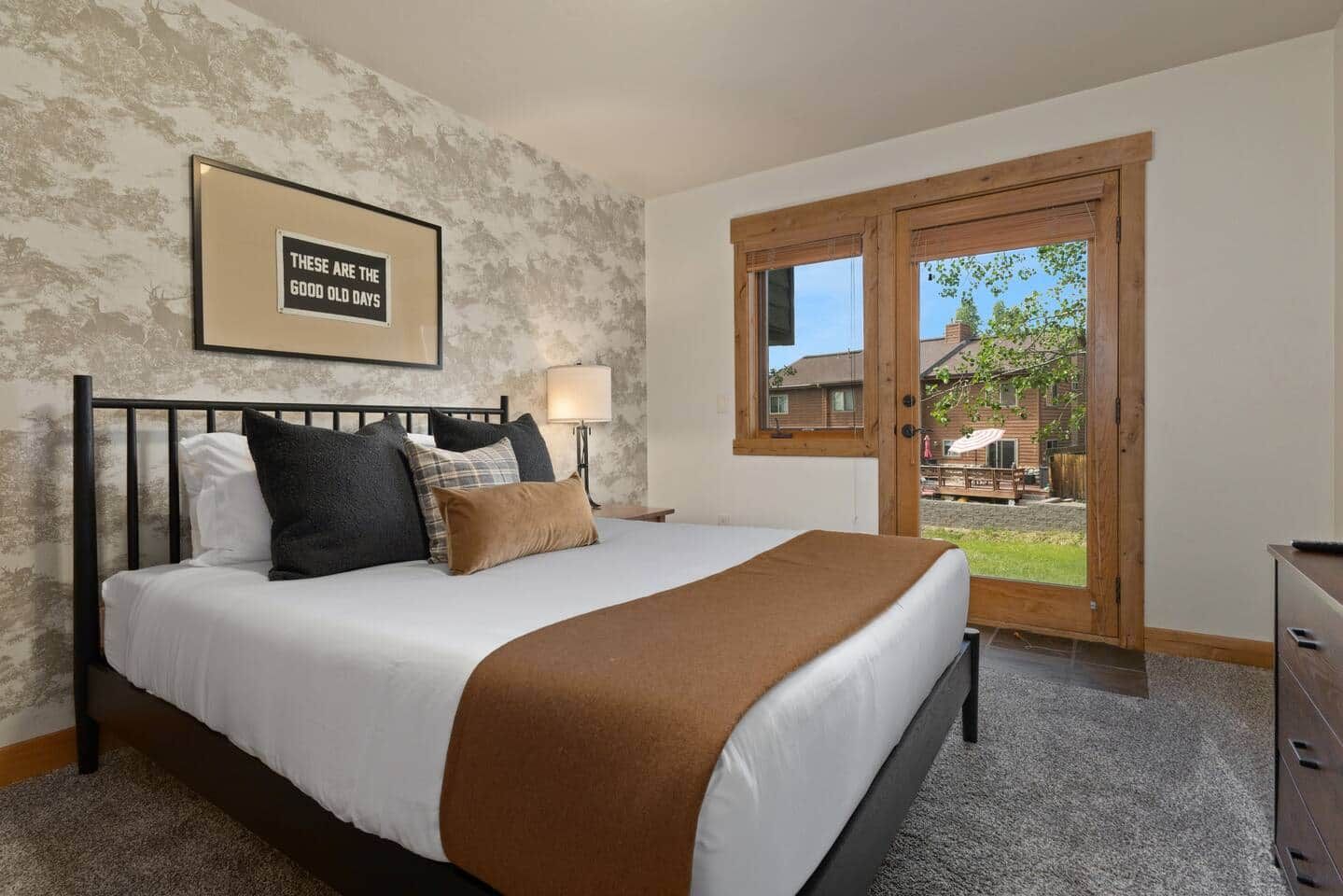 A bedroom with a bed, a nightstand, a lamp, a framed quote on the wall, and a door leading to an outdoor area.