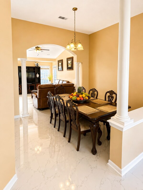A dining area with a marble floor, a wooden dining table with chairs, a fruit bowl, and a ceiling fan.