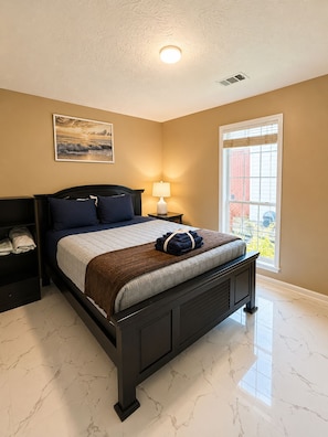 A bedroom with a bed, a nightstand, a lamp, a window, and a painting on the wall.