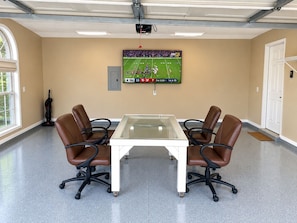 A conference room with a glass-top table, four leather chairs, a mounted TV displaying a sports game, and a vacuum cleaner in the corner.