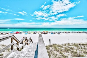 Ein Strand mit einer Holztreppe, die zum Sand hinabführt, ein klarer blauer Himmel und das Meer im Hintergrund.