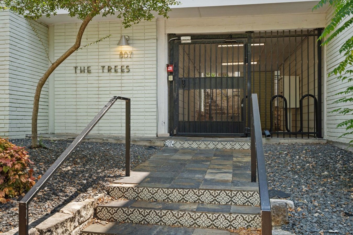 A building entrance with a metal gate, a staircase with patterned tiles, and a tree in the foreground.