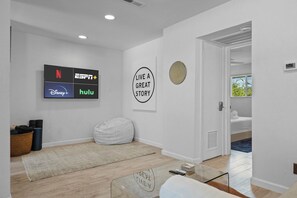 Ein modernes Wohnzimmer mit einem Flachbildfernseher, der Logos von Streaming-Diensten zeigt, einem Sitzsack, einem Motivationsplakat und einem Glastisch.