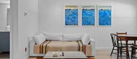 A modern living room with a sofa, a glass coffee table, and a dining table with chairs. There are three abstract paintings on the wall.