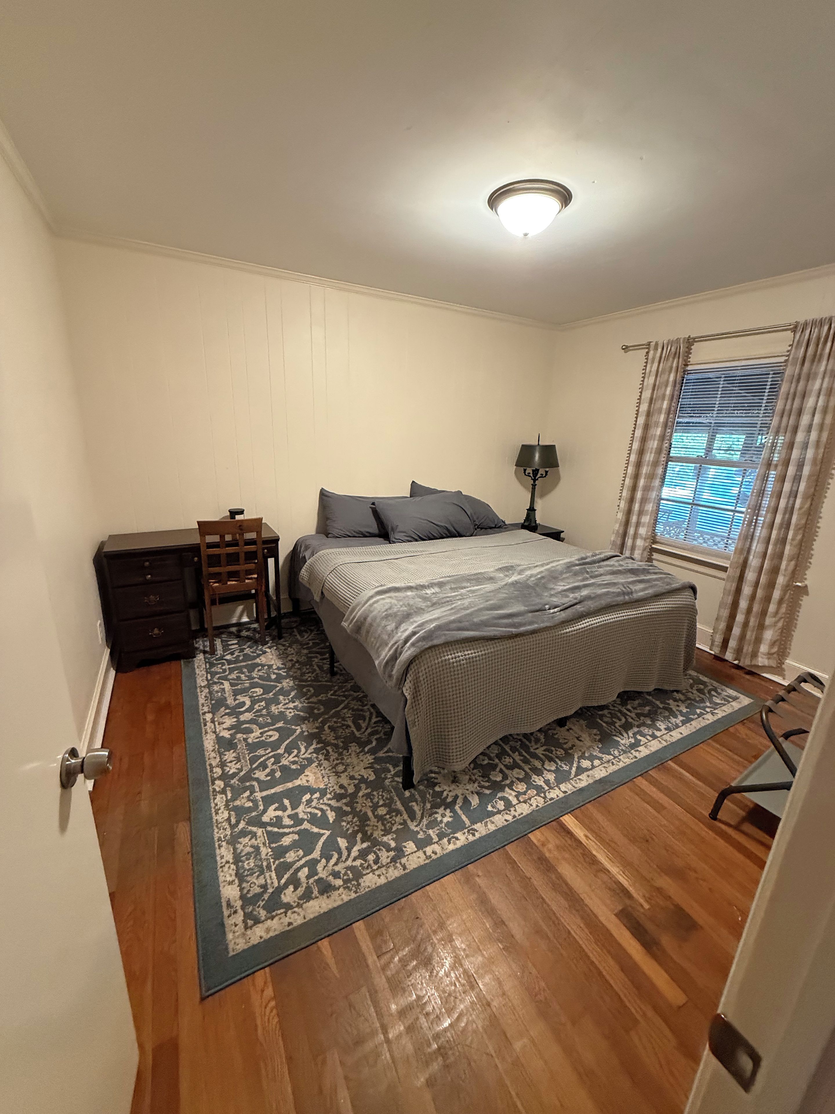 A bedroom with a bed, a nightstand, a lamp, a dresser, and a window with curtains.
