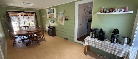A dining area with a wooden table and chairs, a small trash bin, and a coffee station with multiple coffee makers and a kettle.