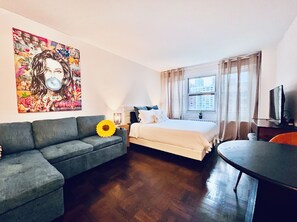 A hotel room with a bed, a sofa, a small table, a TV, and a window with a city view.