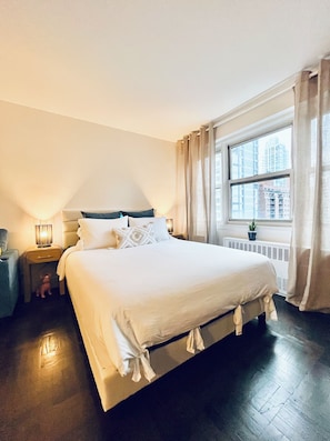 A hotel room with a large bed, a nightstand with a lamp, a window with curtains, and a view of buildings outside. - Midtown East Oversized Studio (New York)