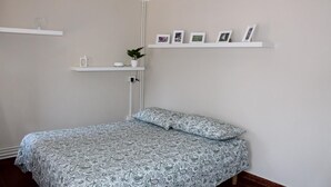 A bedroom with a bed, a nightstand, a shelf with pictures, and a potted plant.