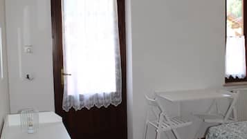 Ein kleines, ordentliches Zimmer mit einem Bett, einem Tisch und zwei Stühlen. Es gibt ein Fenster mit Vorhang, eine Tür und eine Heizung.