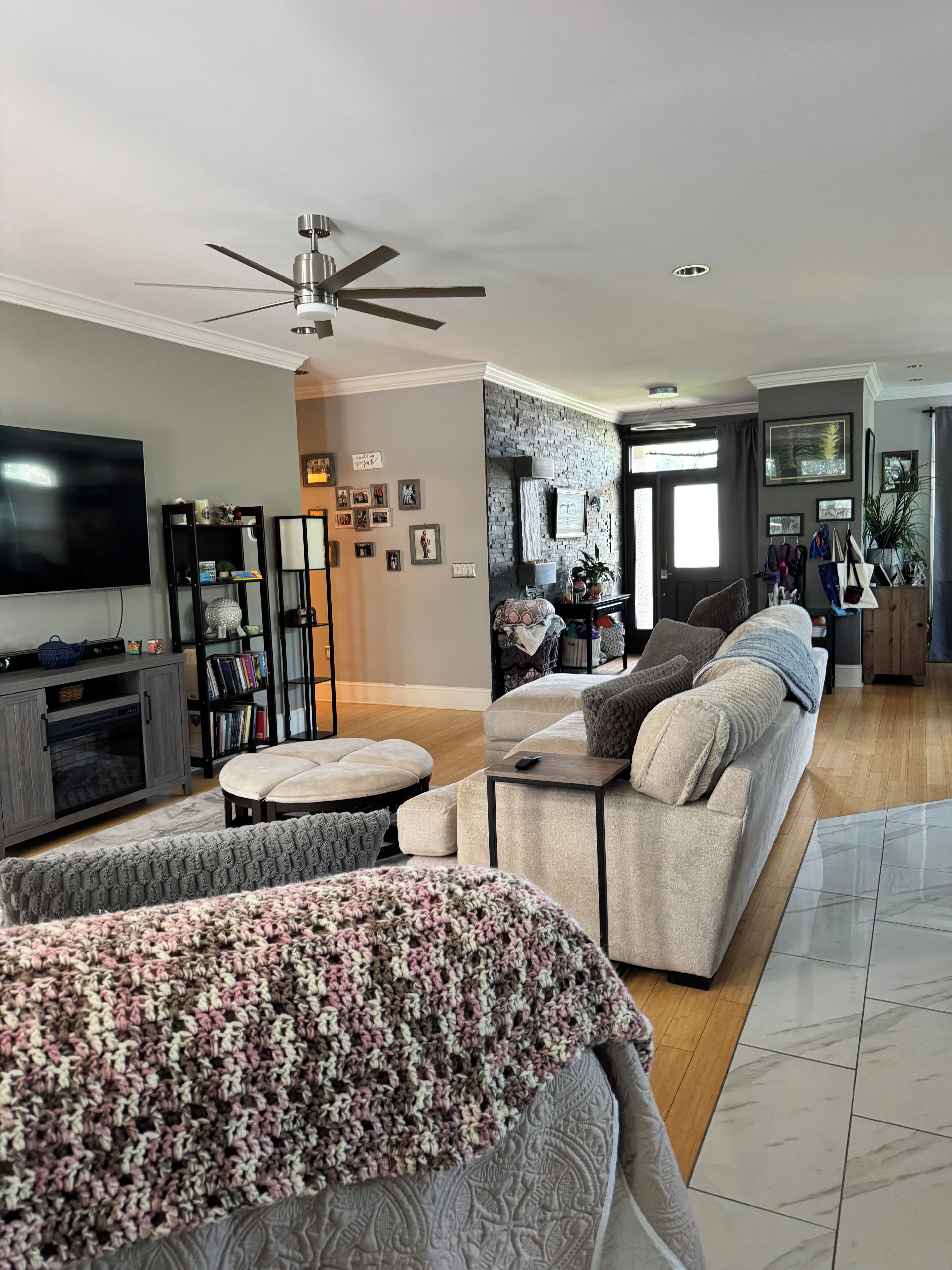 A modern living room with a large flat-screen TV, a ceiling fan, a fireplace, and a cozy seating area with a patterned throw blanket.