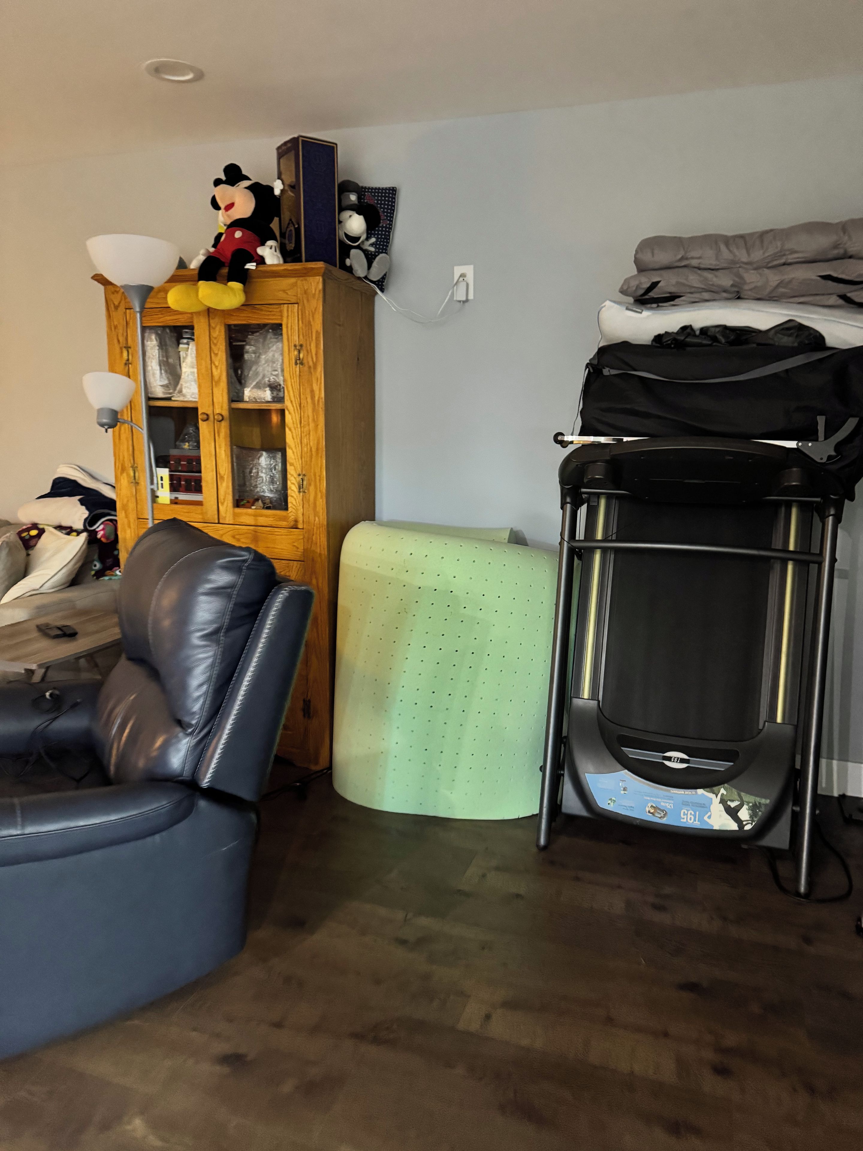 A room with a leather chair, a green ottoman, a wooden cabinet with glass doors, and a black luggage trolley.