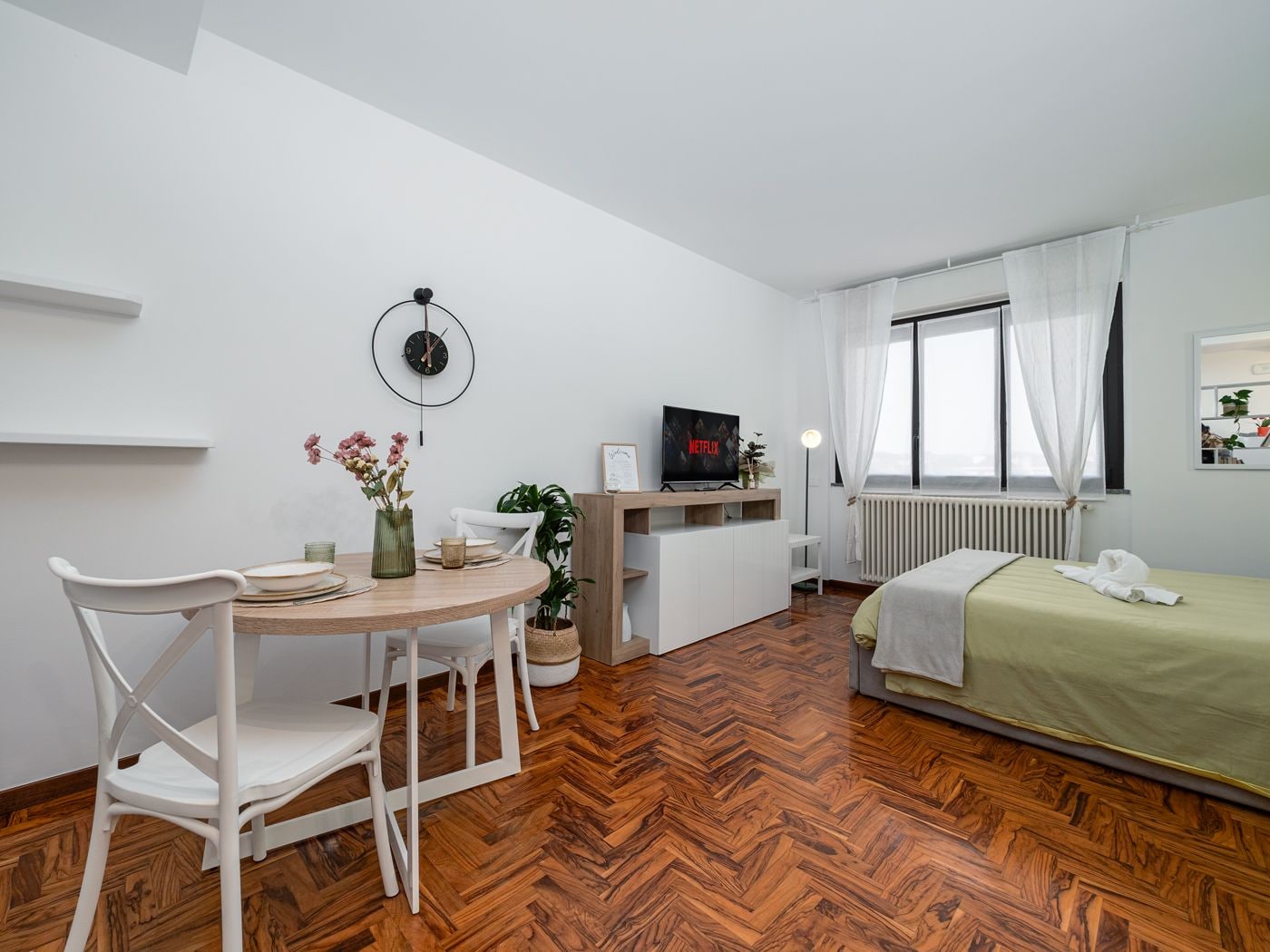 A compact room with a wooden herringbone floor, a bed with a green bedspread, a white chair, a small round table with a vase of flowers, a wall-mounted clock, and a television on a stand.