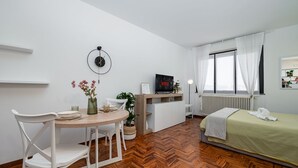 Una camera compatta con pavimento in legno a spina di pesce, un letto con copriletto verde, una sedia bianca, un tavolino rotondo con un vaso di fiori, una sveglia a parete e un televisore su un mobile.