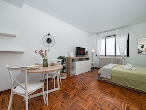 Een knus ingerichte kamer met een houten visgraatvloer, een bed met een groene plaid, een witte stoel, een klein rond tafeltje met een bloevas, een wandklok en een televisie op een dressoir.