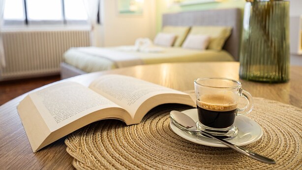Una stanza con un letto, un comodino con un libro e una tazza di caffè, e un vaso con dei fiori.