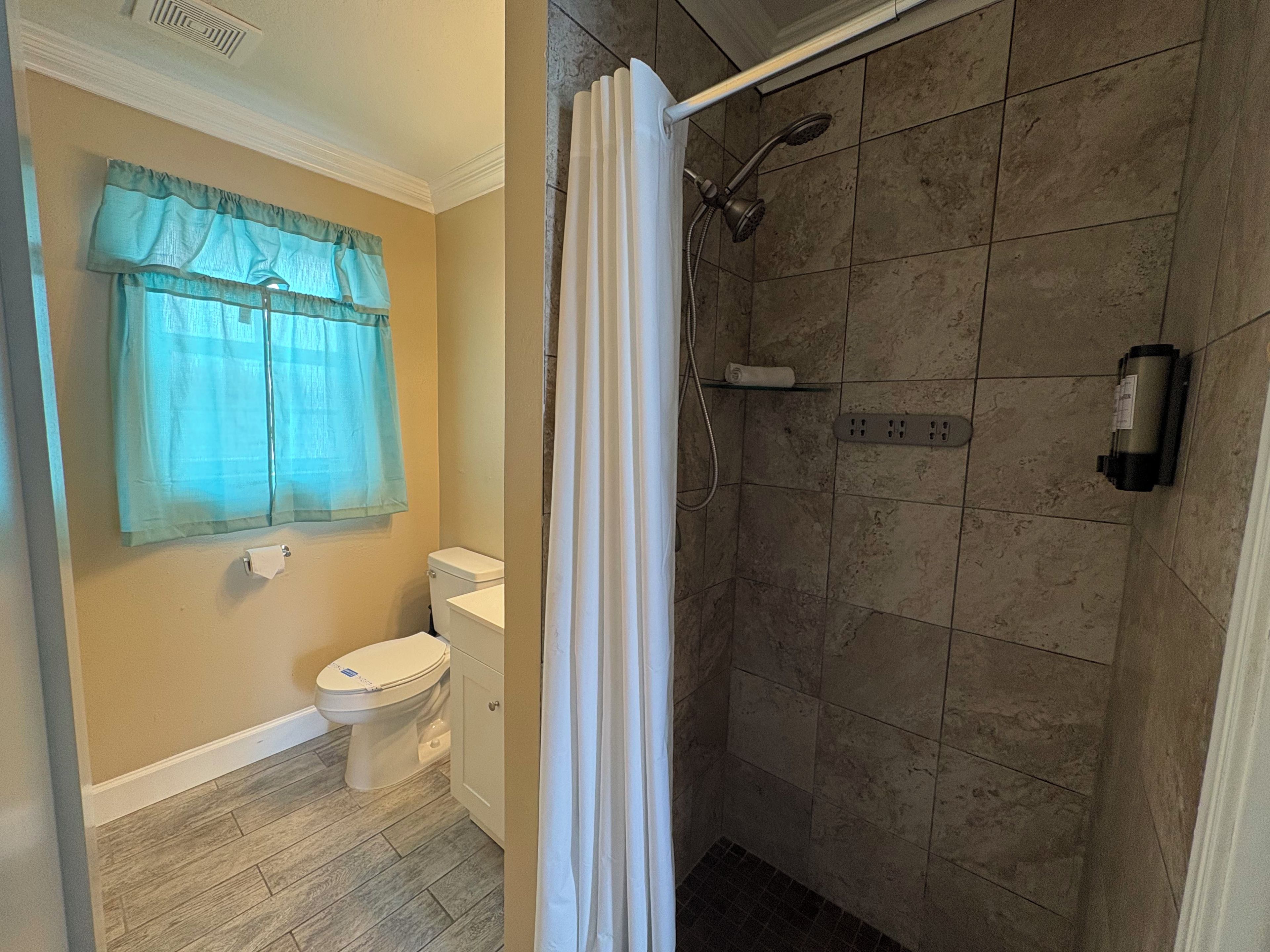 A bathroom with a toilet, a shower with a glass door, and a window with a curtain.