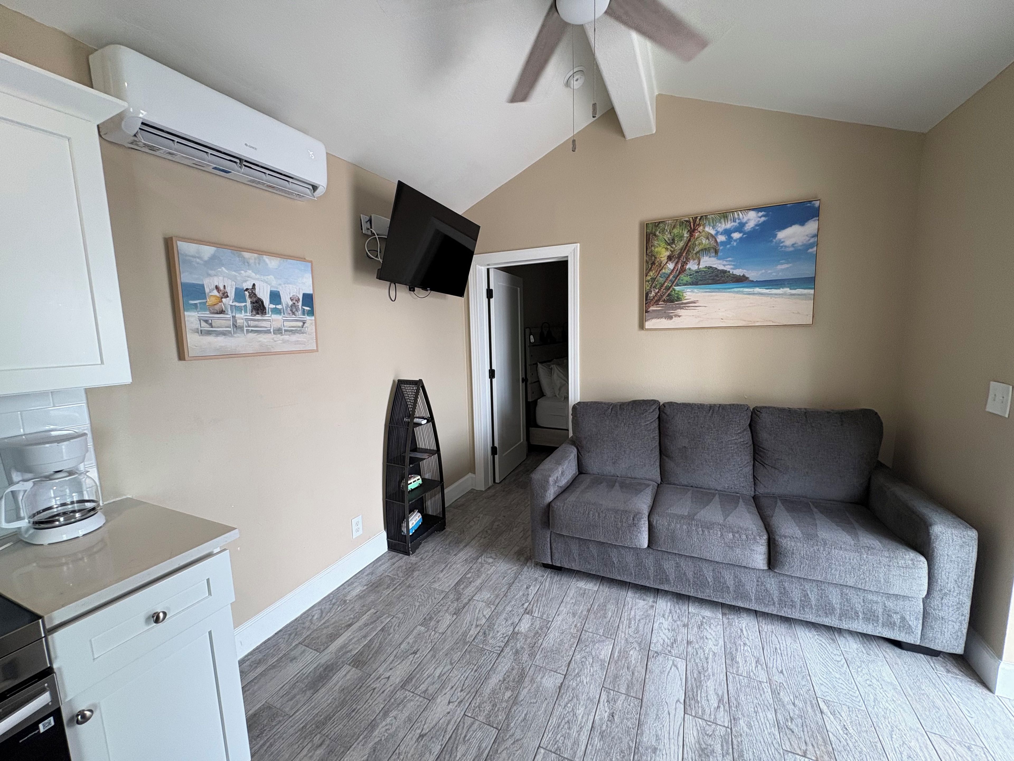 Un salon avec un canapé gris, un ventilateur de plafond, un téléviseur à écran plat, un tableau représentant une scène de plage et une machine à café.