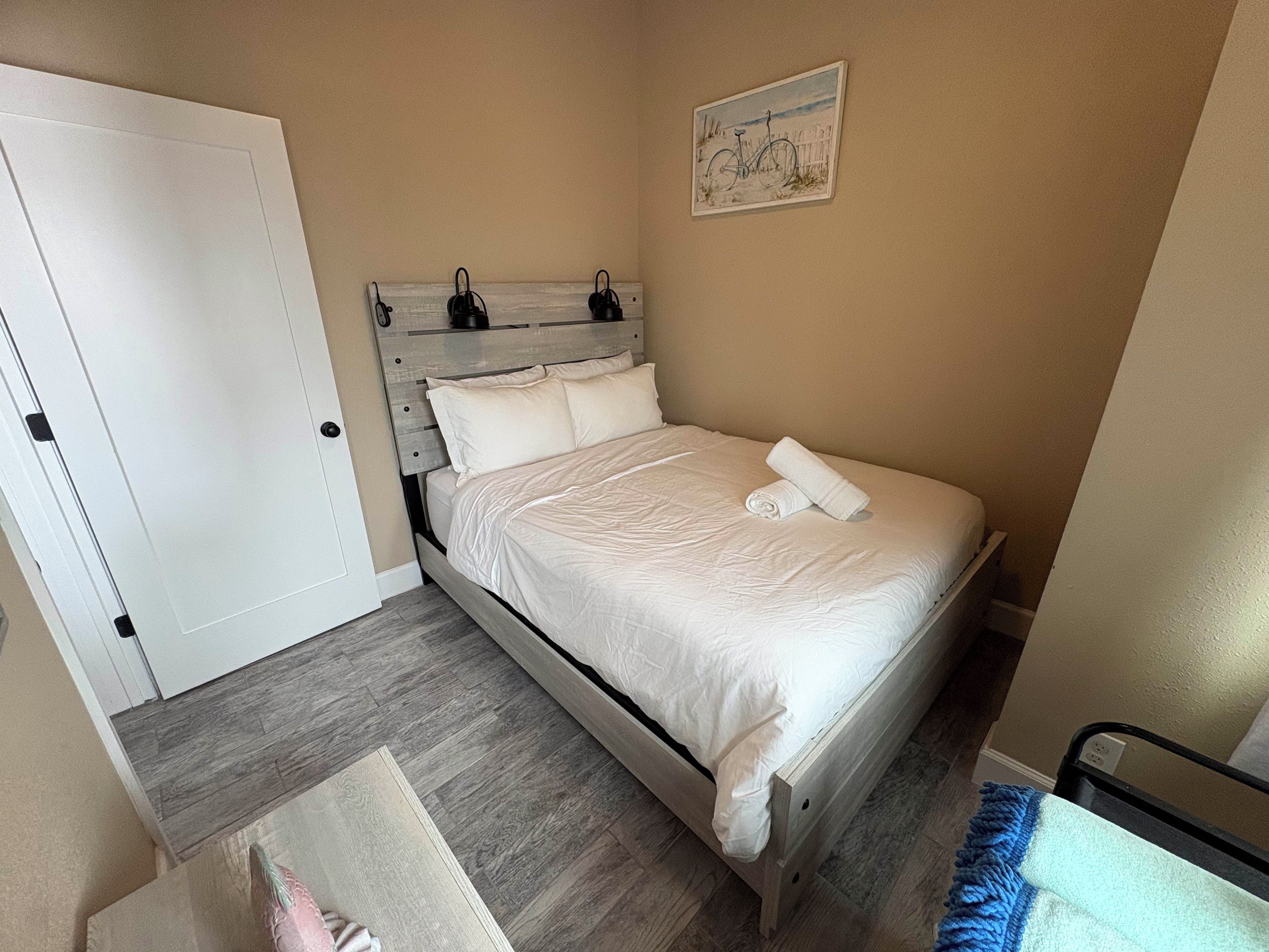 A bedroom with a wooden bed, white bedding, a painting on the wall, and a door.