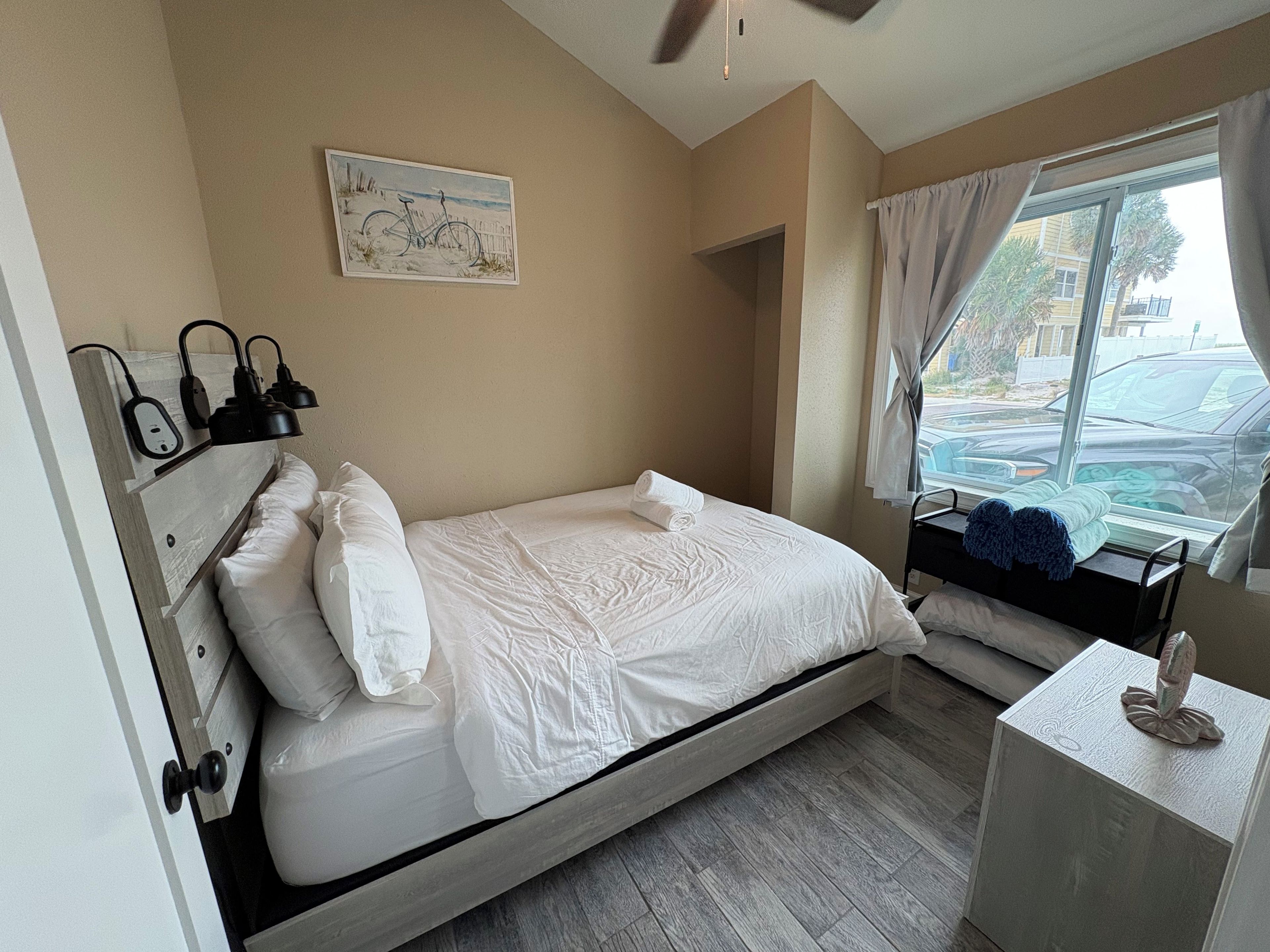 A single-bed room with a wooden headboard, a bedside table with a lamp, a window with curtains, and a painting on the wall.