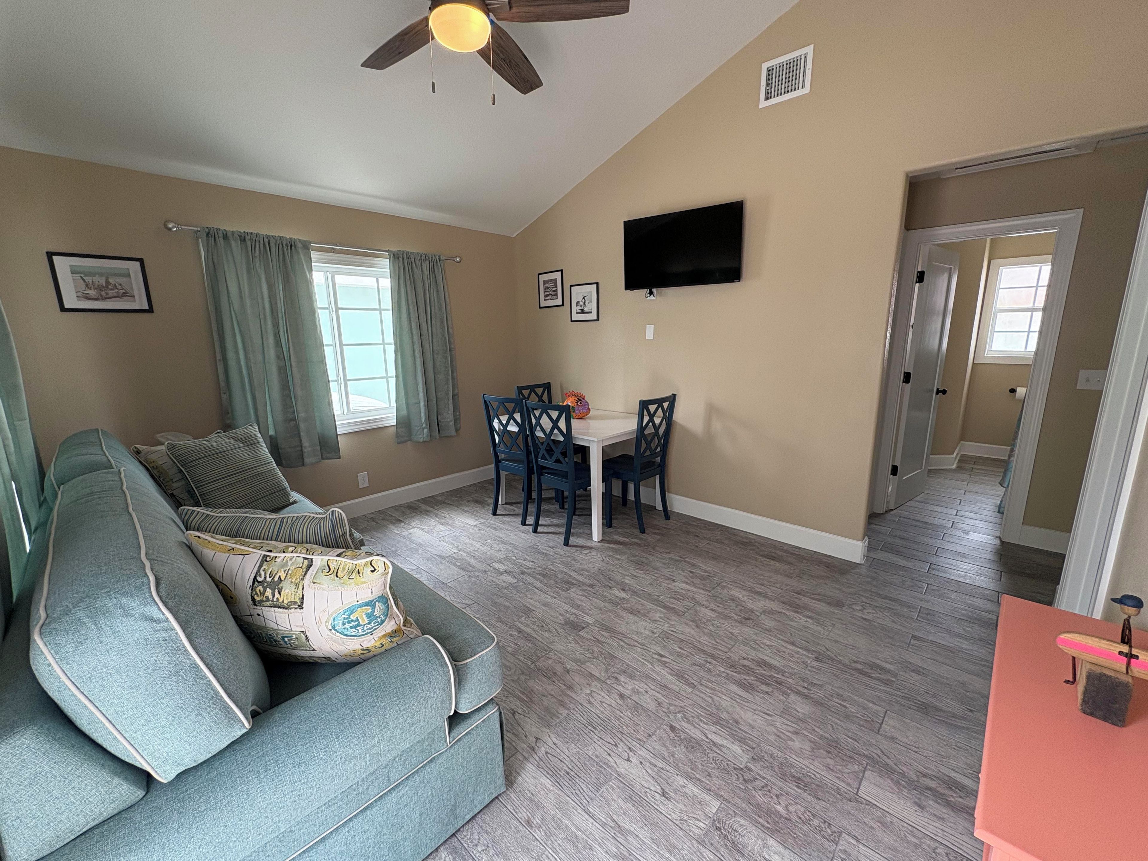 A living room with a sofa, dining table, chairs, a ceiling fan, and a TV mounted on the wall.