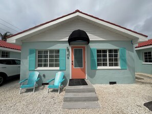 A two-story building with a red roof, teal shutters, and a black awning. There are two blue Adirondack chairs outside. - 🏖️ 2 BR Beach Bungalow I Unit 10 (Treasure Island)