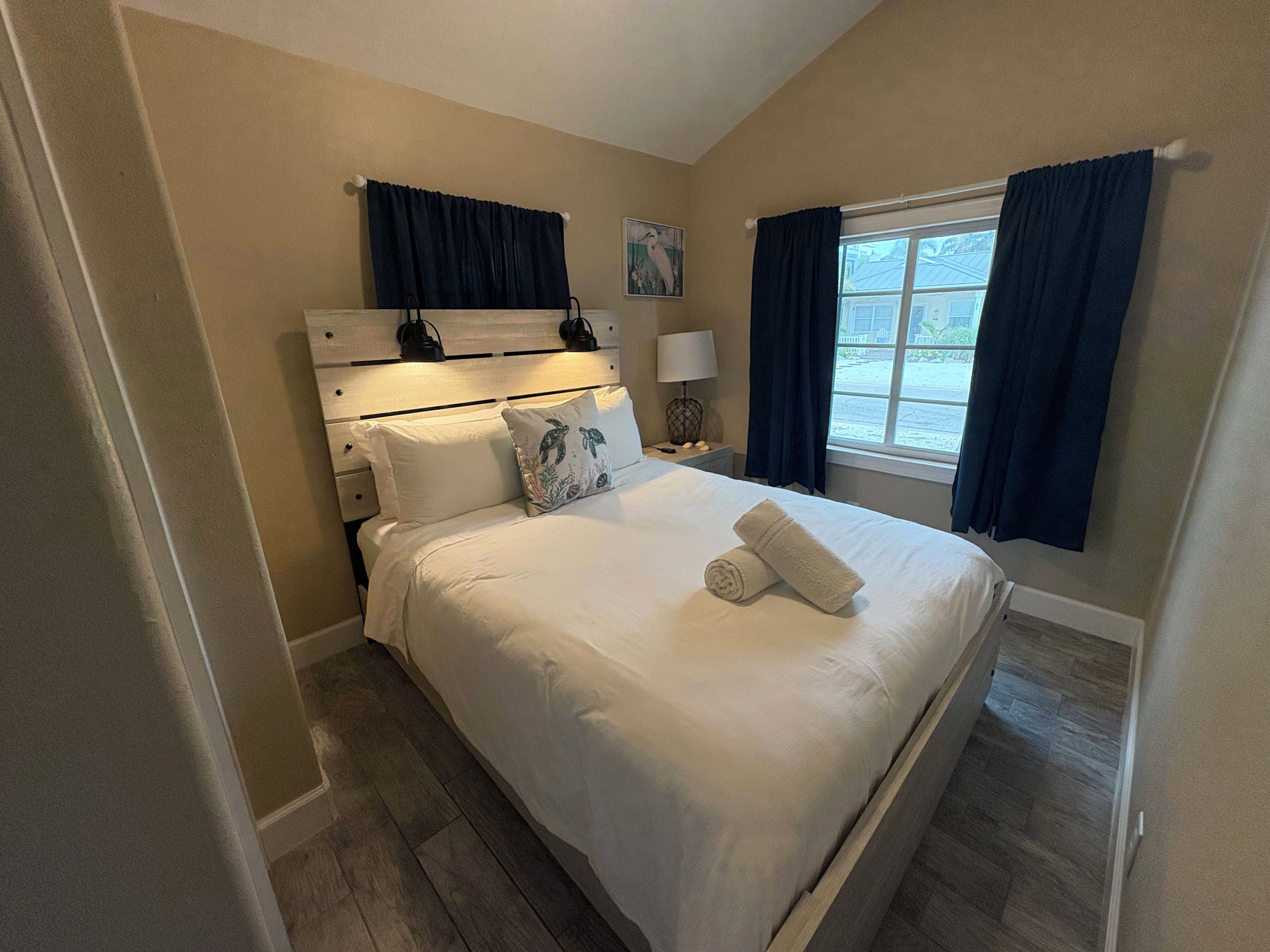 A neatly made bed with white linens and a rolled towel, a bedside lamp, a nightstand with a lamp, and a window with curtains.