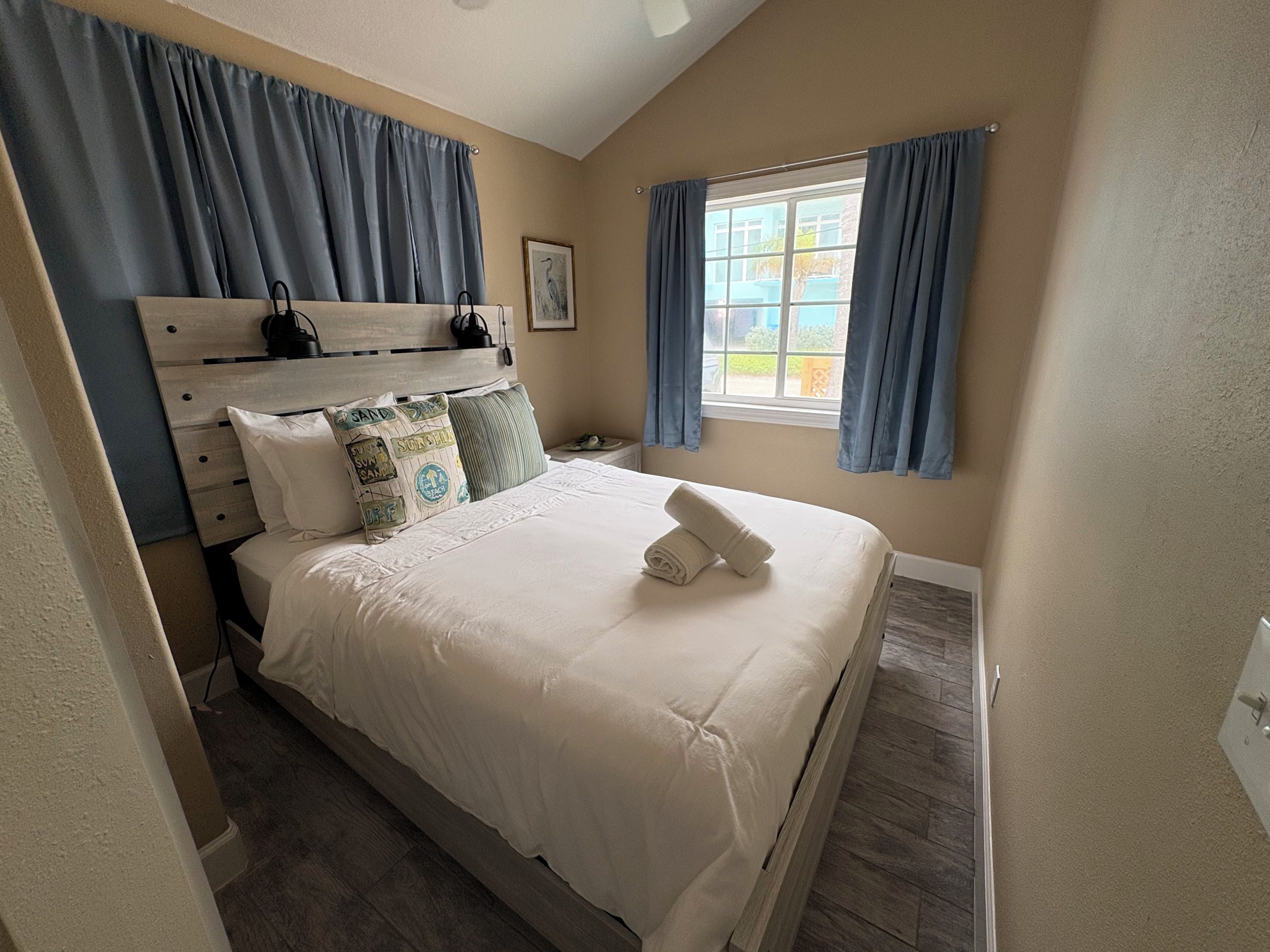 Une chambre à coucher avec un lit, une tête de lit, une fenêtre avec des rideaux, un tableau encadré et une table de chevet avec une lampe.