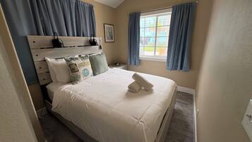 Une chambre à coucher avec un lit, une tête de lit, une fenêtre avec des rideaux, un tableau encadré et une table de chevet avec une lampe.
