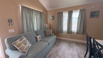 A living room with a sofa, a dining table with chairs, a window with curtains, and a lamp.