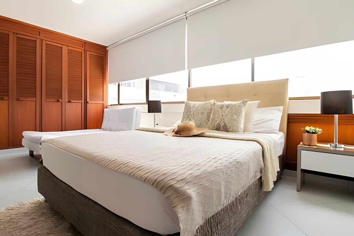 A modern bedroom with a large bed, wooden paneling, a bedside table with a lamp, and a view through the window.