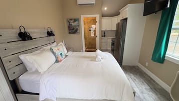A bedroom with a bed, a desk, a chair, a television, and a kitchen area with cabinets and a refrigerator.