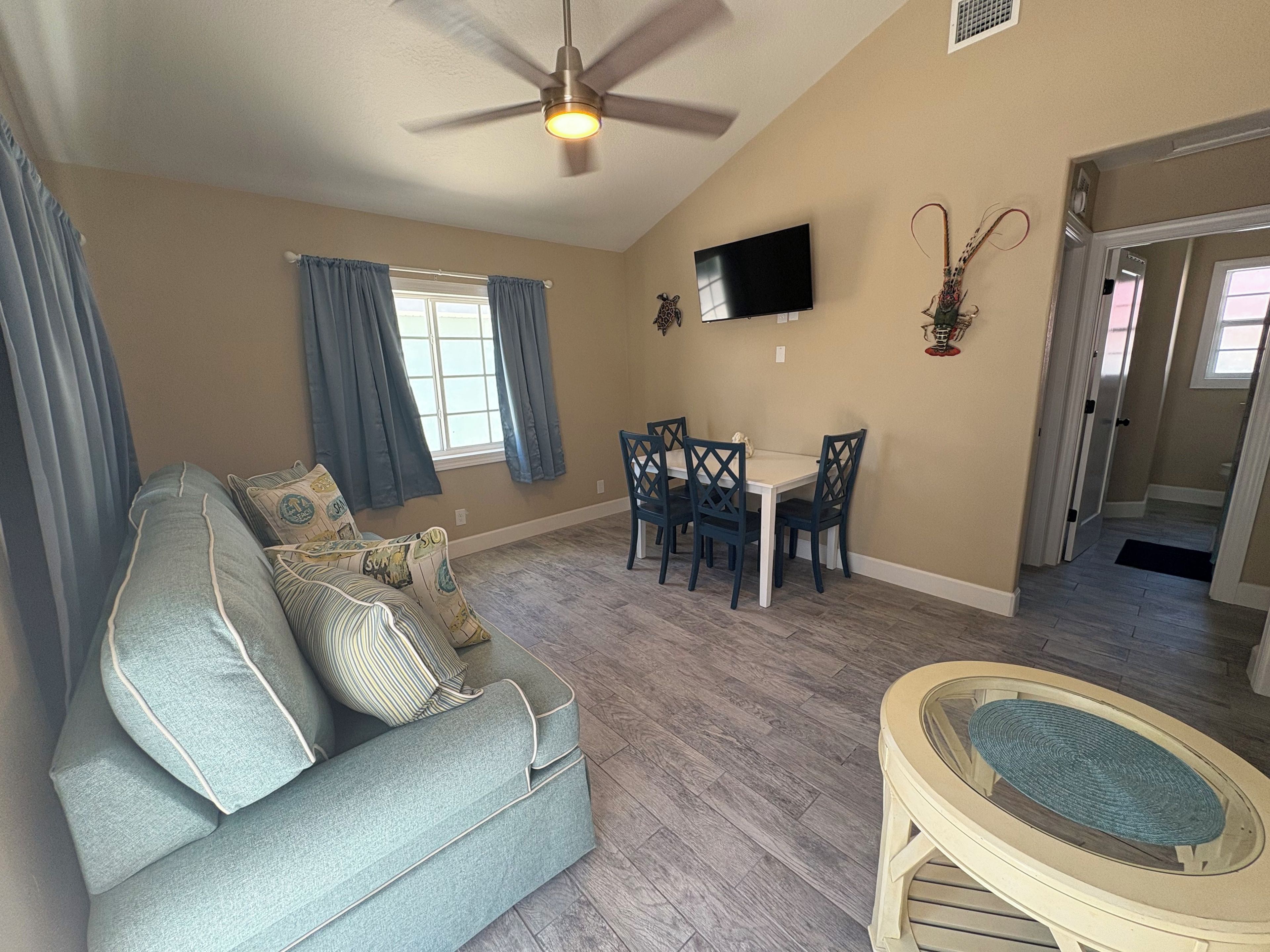 A living room with a sofa, dining table, chairs, a ceiling fan, a flat-screen TV, and a door leading to another room.