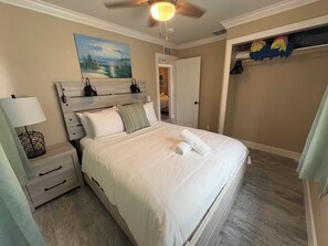 A bedroom with a bed, nightstand, lamp, painting, closet, and a ceiling fan. - 🏡 Beach Bungalow Collection | Sleeps 24 (Treasure Island)