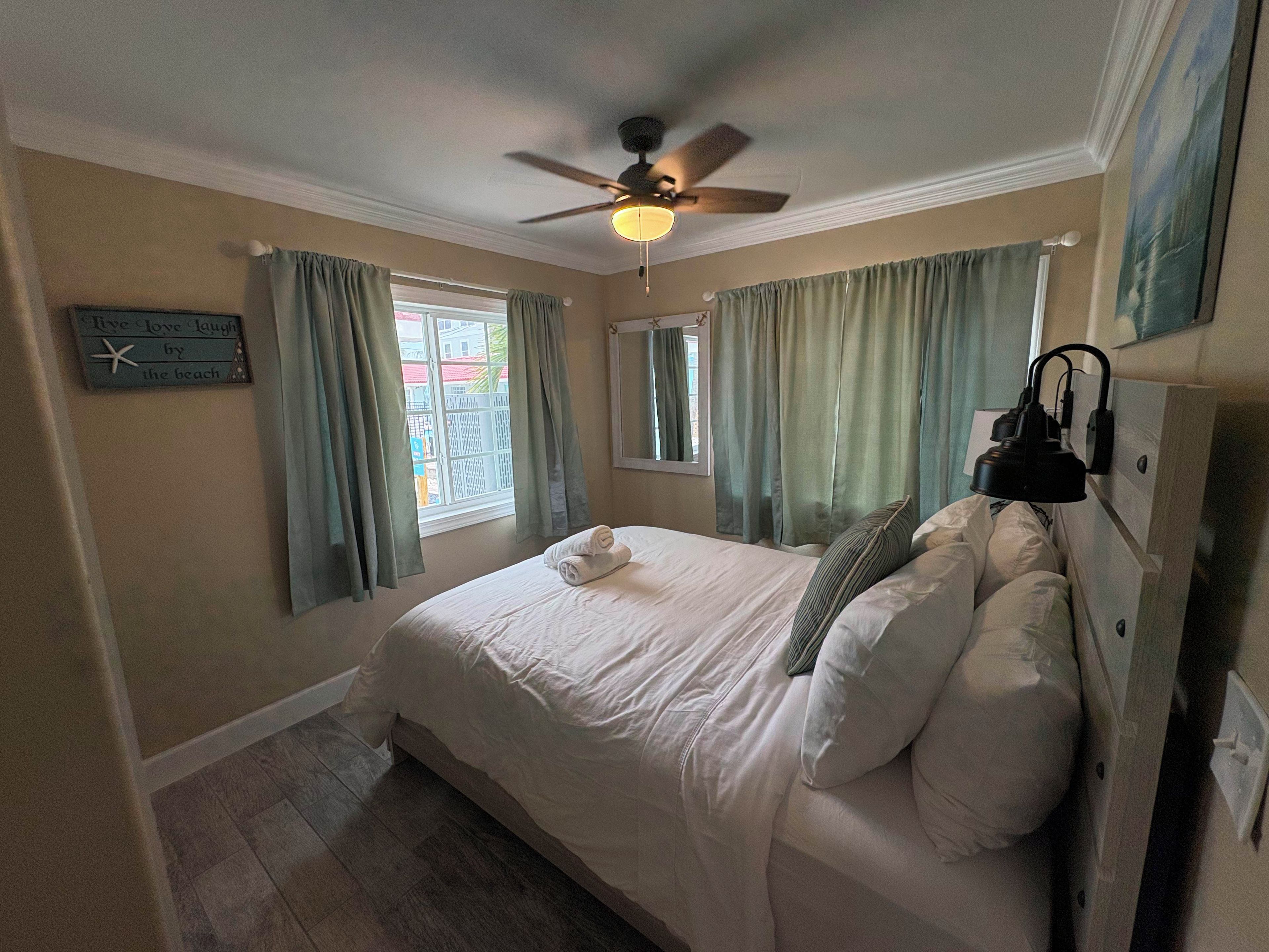 A bedroom with a bed, a ceiling fan, a window with curtains, a wall clock, and a bedside lamp.