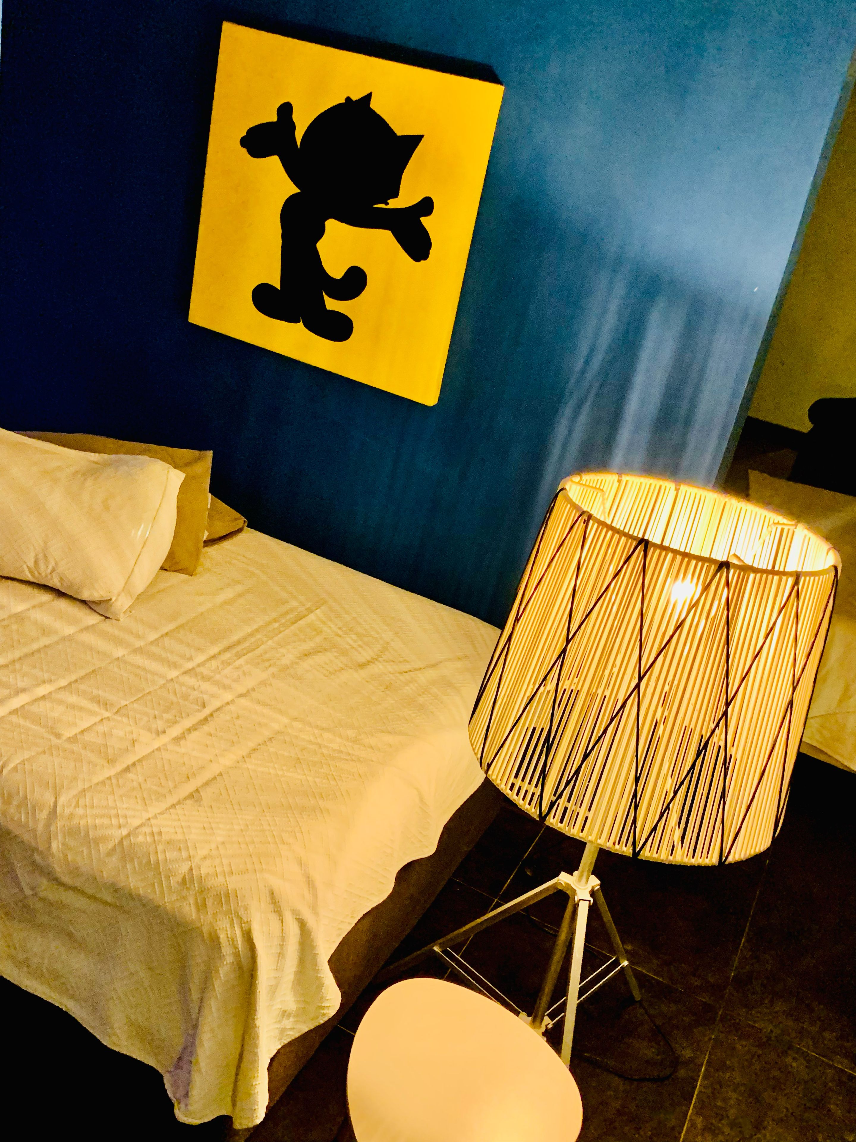 Un lit avec une literie blanche, une lampe avec un abat-jour en osier et un mur orné d’une enseigne jaune sur laquelle figure une silhouette noire.