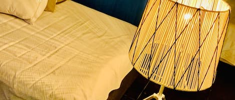 A bed with white bedding, a lamp with a woven shade, and a wall with a yellow sign featuring a black silhouette.