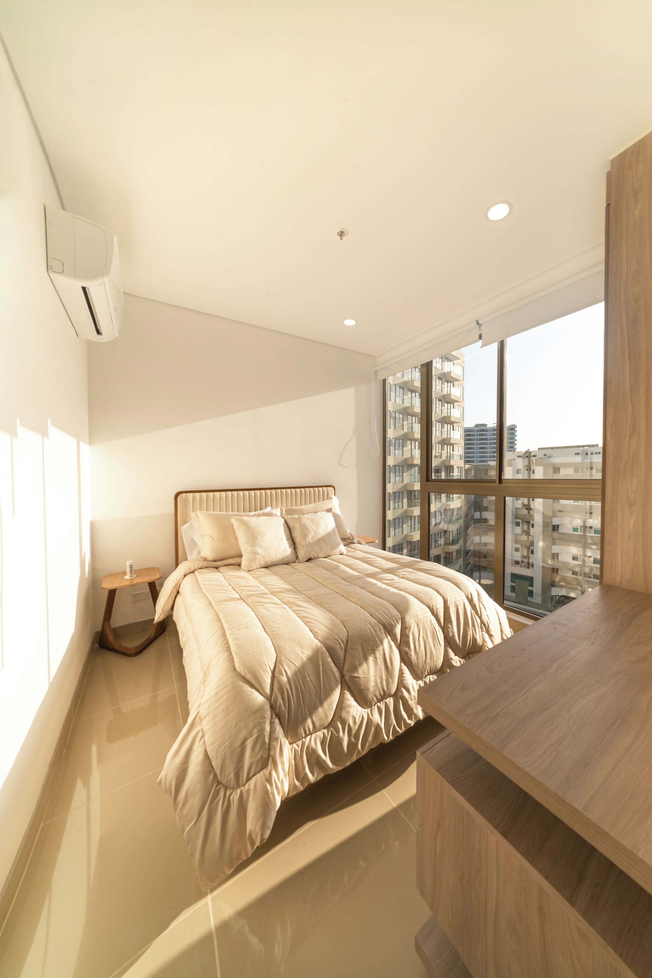 A modern bedroom with a large bed, a bedside table, a wooden desk, and a view of high-rise buildings.
