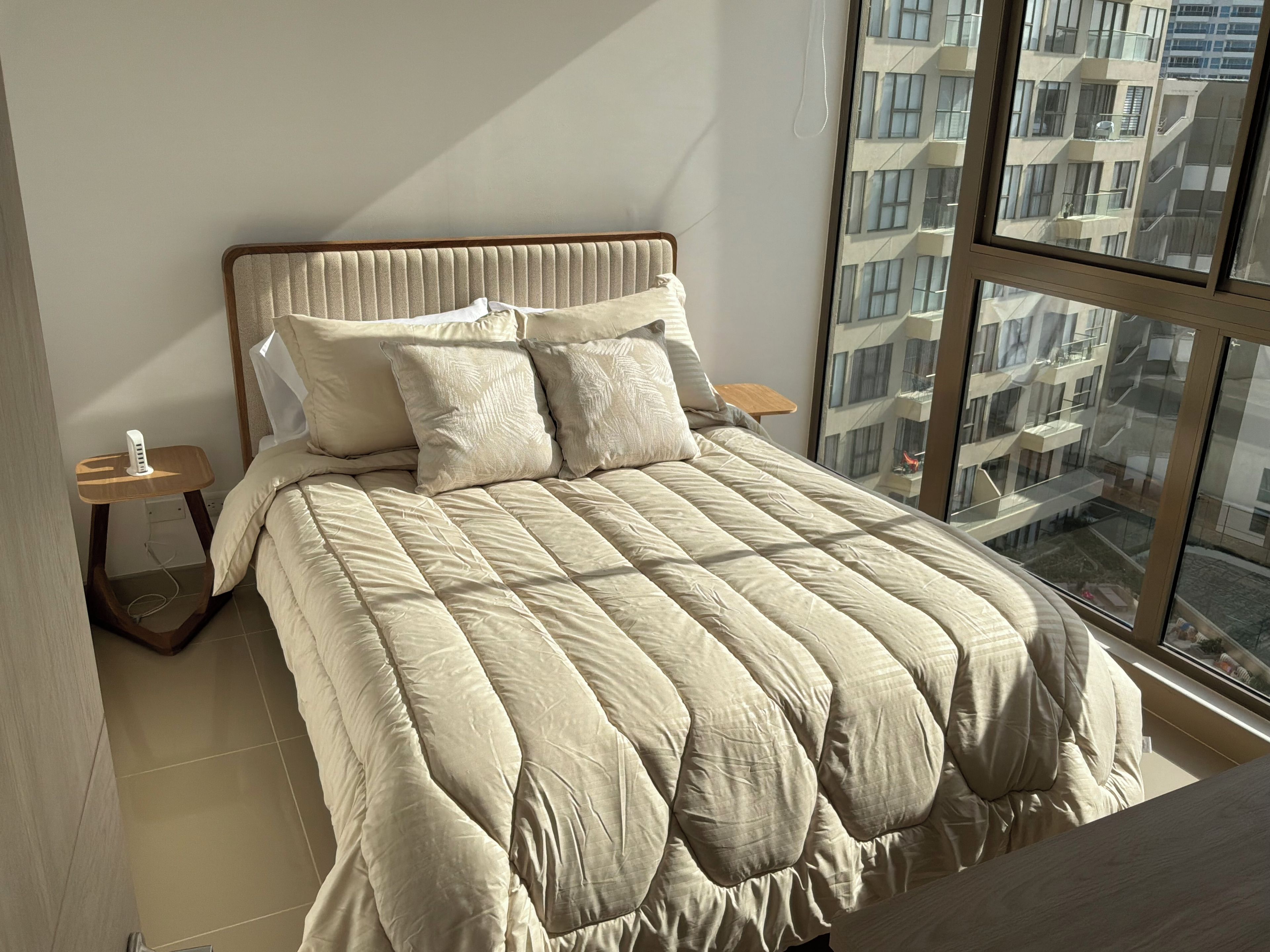 A bedroom with a large bed, a bedside table, a nightstand, and a window with a view of buildings.