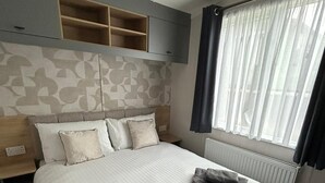 Een netjes opgemaakt bed met kussens en handdoeken, een hoofdbord met ingebouwde planken, een nachtkastje en een raam met gordijnen.