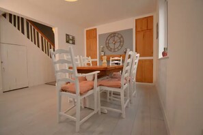 Un coin repas avec une table en bois et des chaises blanches, une horloge au mur et un escalier en arrière-plan.
