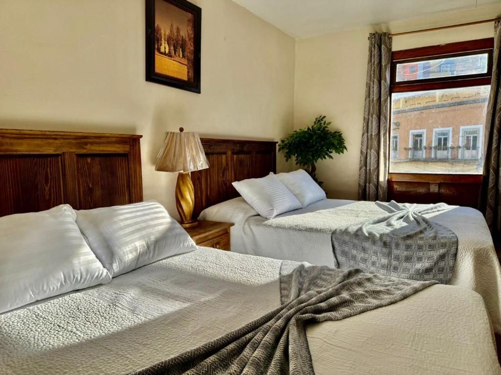 A hotel room with two beds, a nightstand, a lamp, a plant, and a window with a view of buildings.
