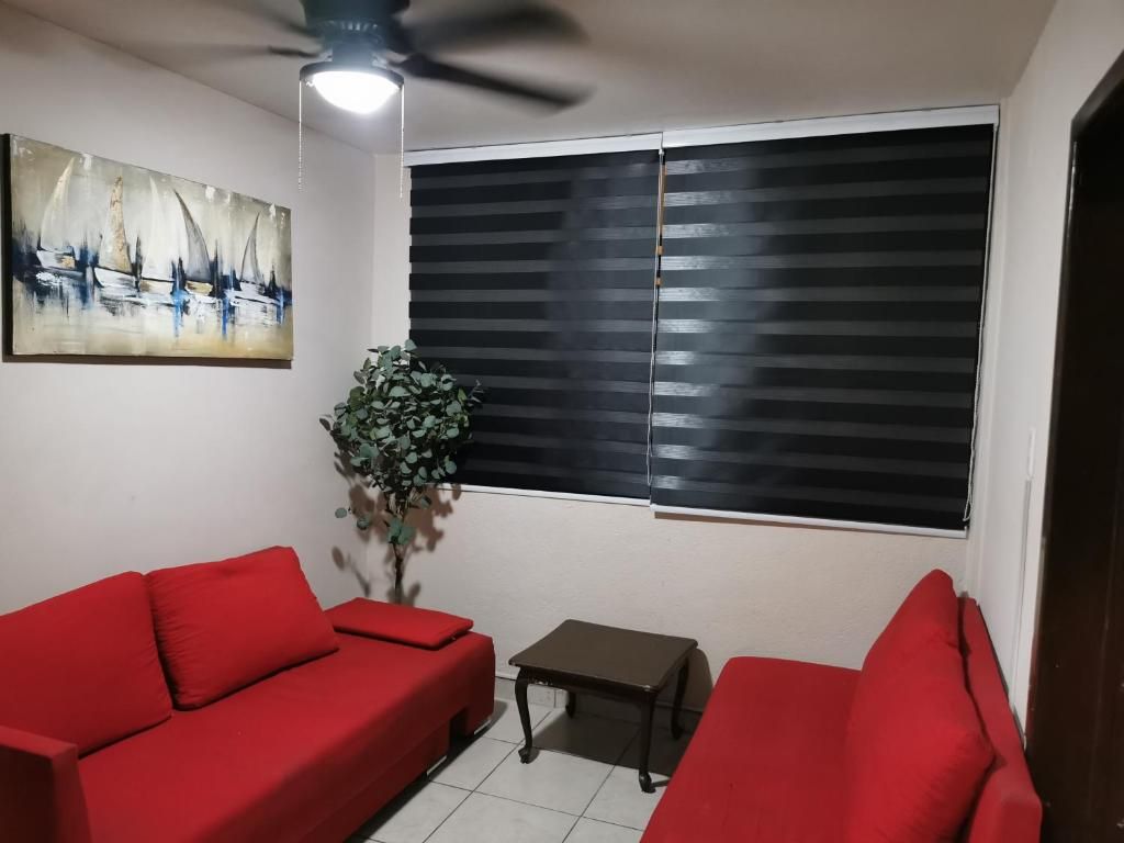 Una sala de estar con dos sofás rojos, una mesita, un ventilador de techo, un cuadro y una ventana con persianas.