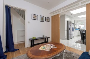 Una sala moderna con una mesa de centro de madera, una revista y un bol de manzanas. Se ve una escalera que conduce a otra habitación, y un área de cocina al fondo.