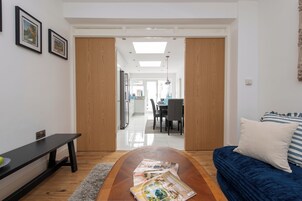 Uma sala de estar moderna com sofá, mesa de centro com revistas, um banco e uma área de refeições ao fundo.