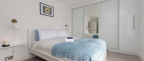 A modern bedroom with a bed, bedside lamps, a bedside table, a wall-mounted picture, and a large mirror.