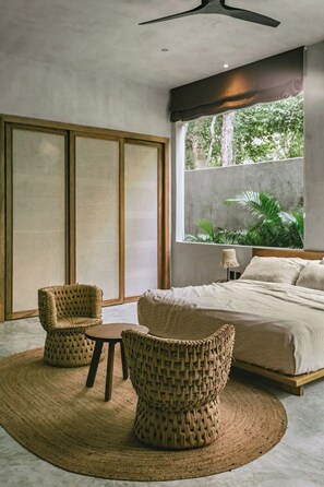 A bedroom with a bed, two wicker chairs, a small table, a ceiling fan, and a large window with a view of greenery. - Casa Shalva with Chef & Transportation Included. Two Pools. (Tulum)